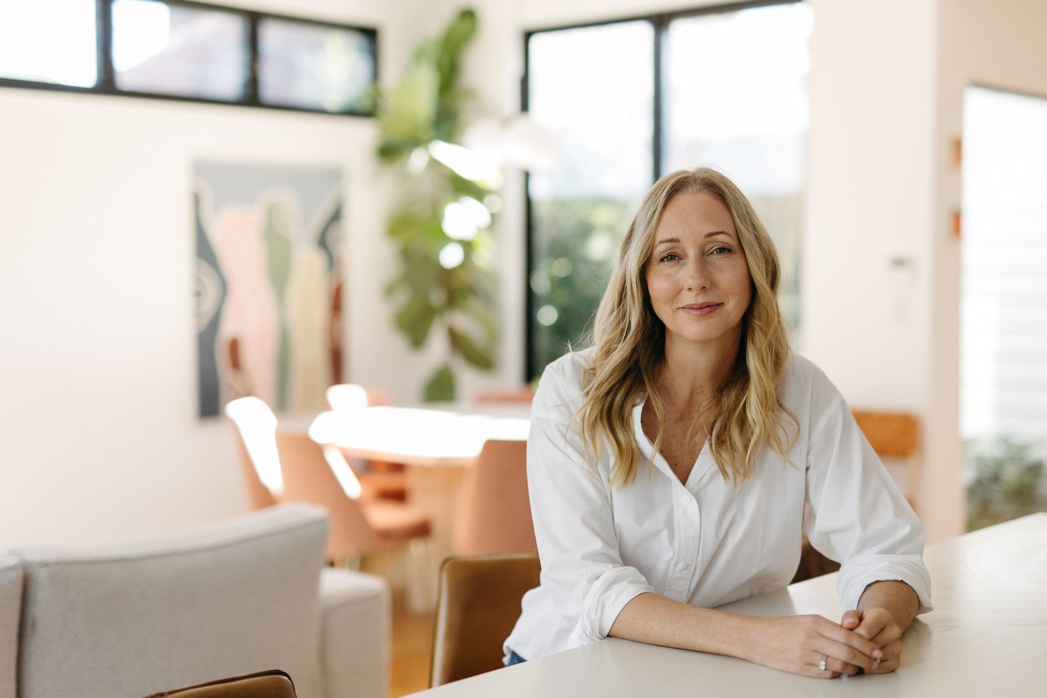 Smiling blonde woman in white shirt sitting at a modern white table in a bright, stylish open-plan living space.