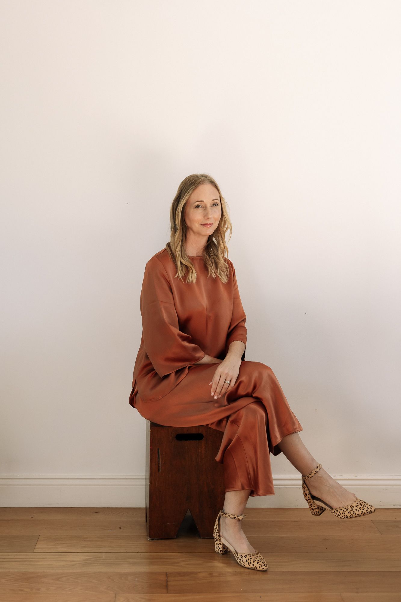 Woman in rust satin midi dress and leopard heels sitting on wooden stool against white wall.