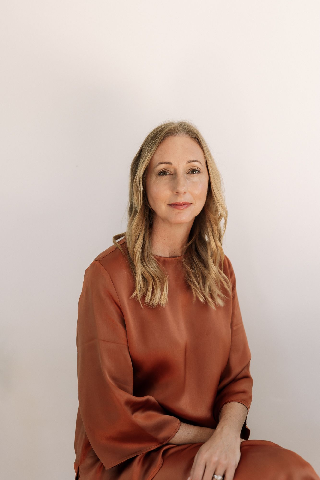 Woman in rust-colored satin blouse seated against white background, with blonde hair and calm expression.
