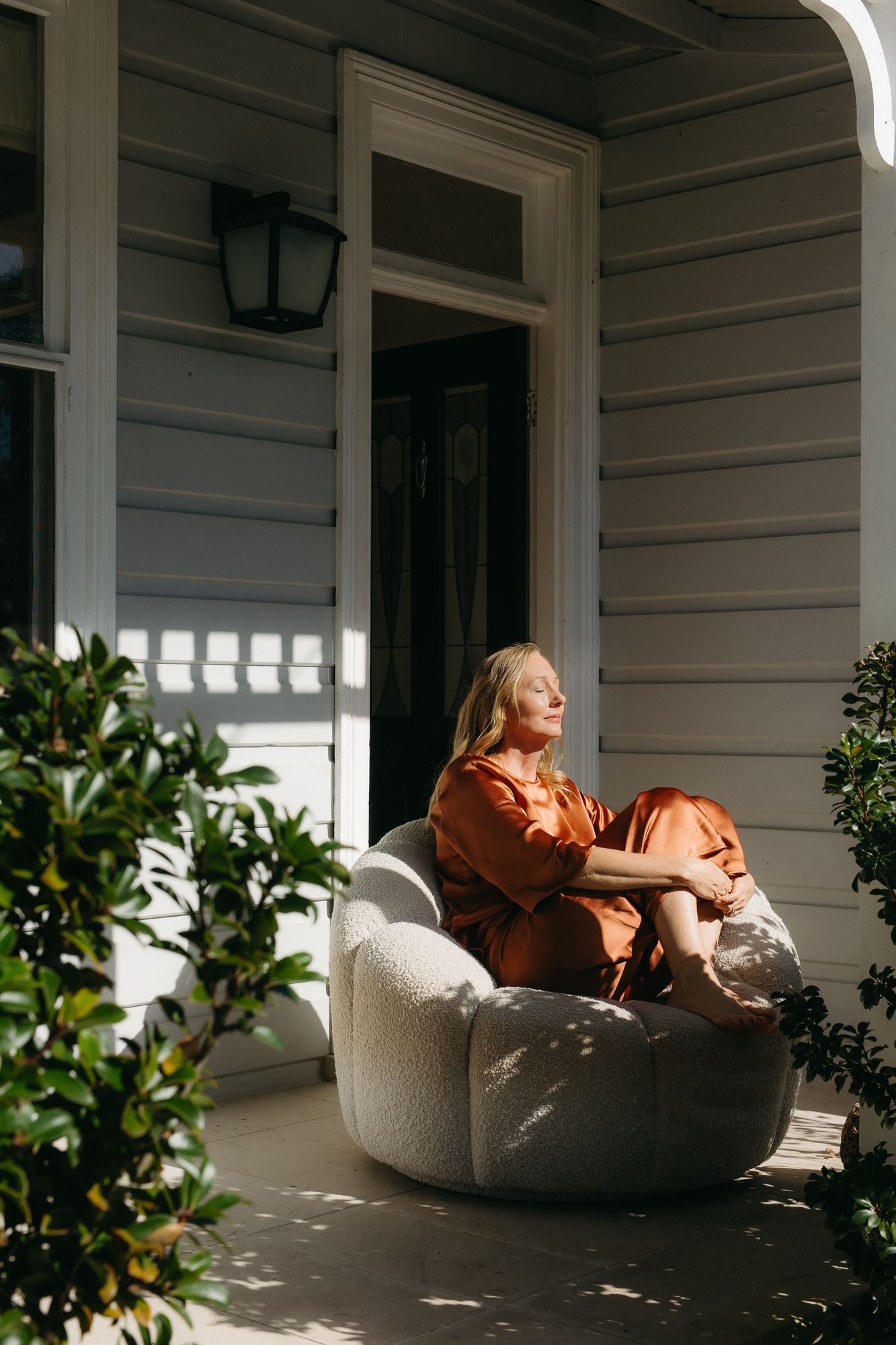 Woman in rust orange outfit relaxing on a round chair on a sunlit porch of a grey weatherboard home.