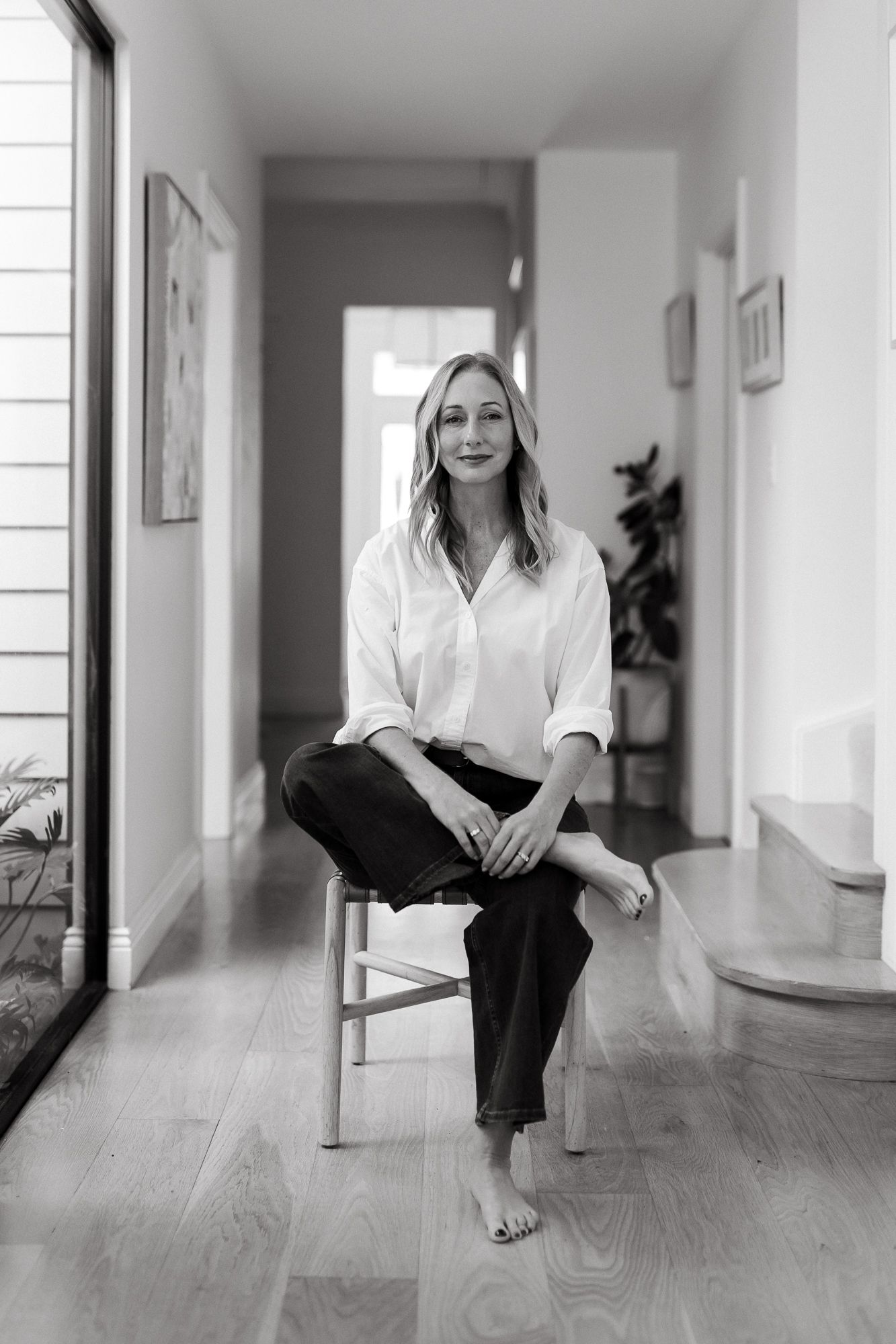 Black and white portrait of a woman in white shirt and dark jeans sitting on a stool in a modern hallway.