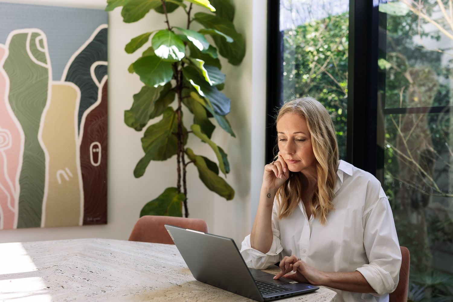Woman in white shirt working on laptop at table with modern art and tropical plant in bright home office.