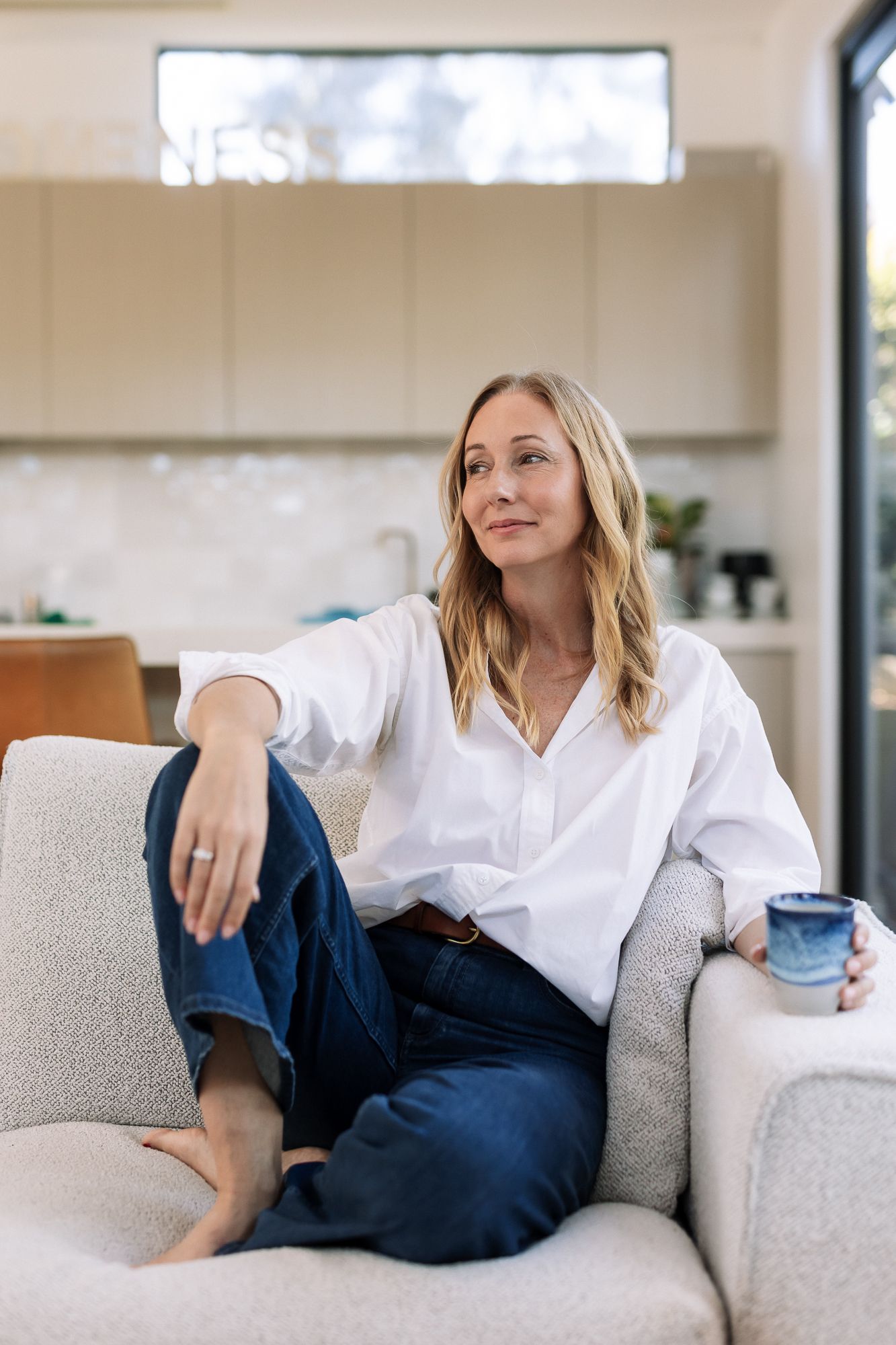 Woman in white shirt and jeans relaxing on sofa with blue mug in modern living room.