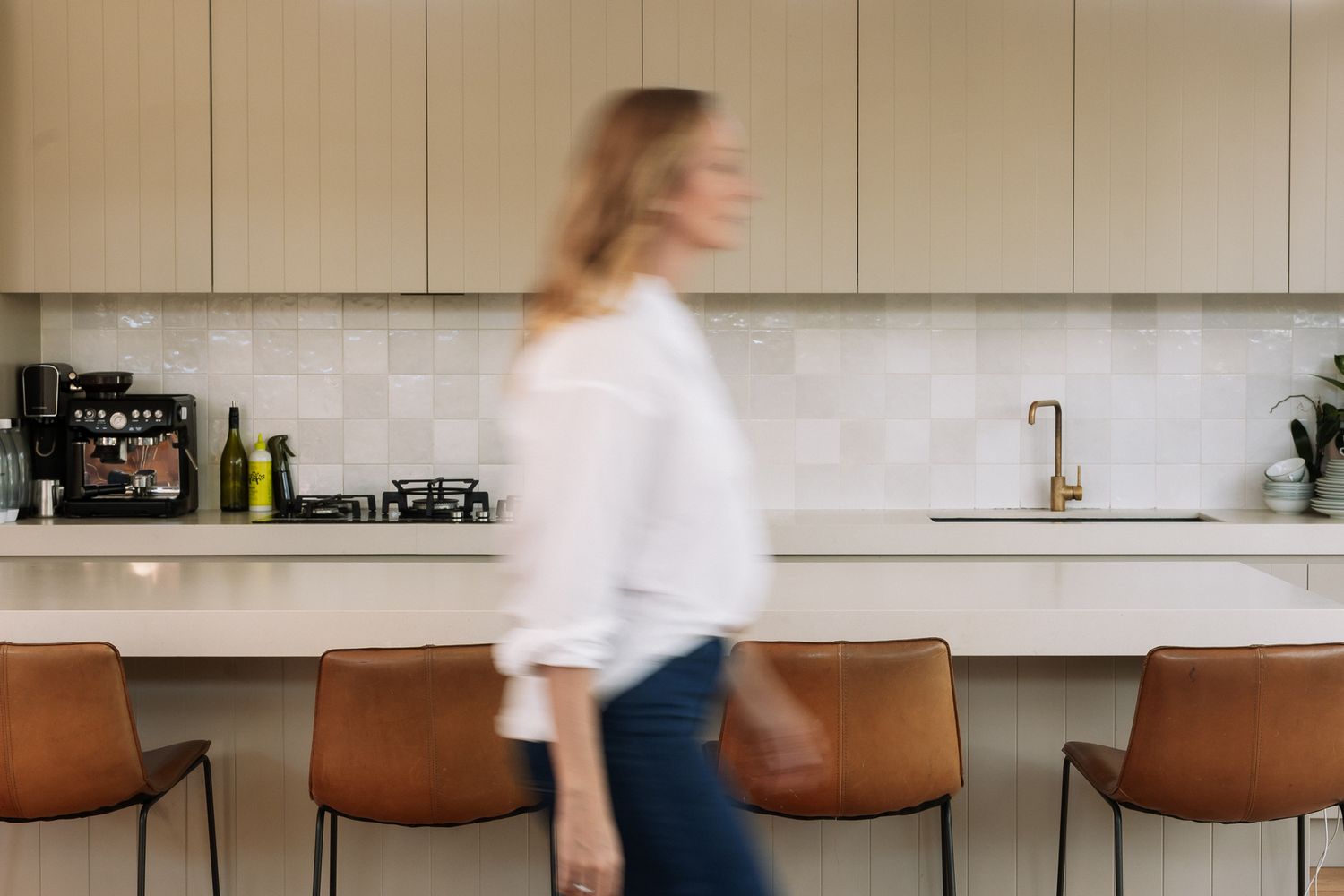 Woman walking through a modern kitchen with beige cabinetry, white tile backsplash, brass fixtures, and leather bar stools.