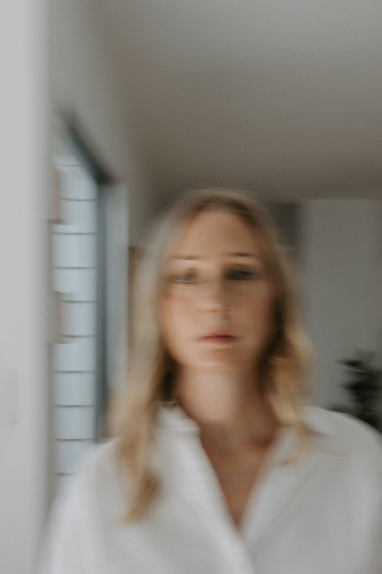 Blurred portrait of a blonde woman in a white shirt standing in a bright minimalist interior space.