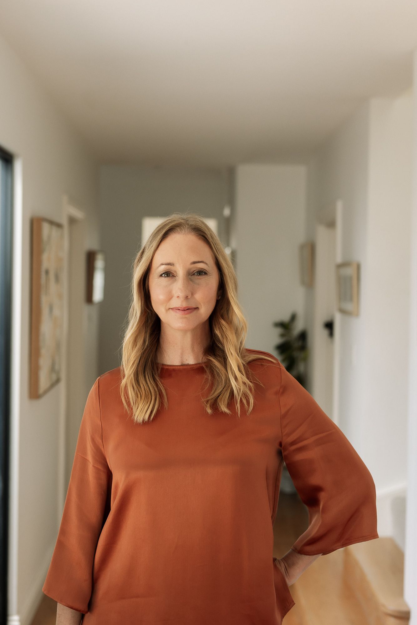 A woman in a rust-colored blouse stands confidently in a modern home hallway with neutral decor.
