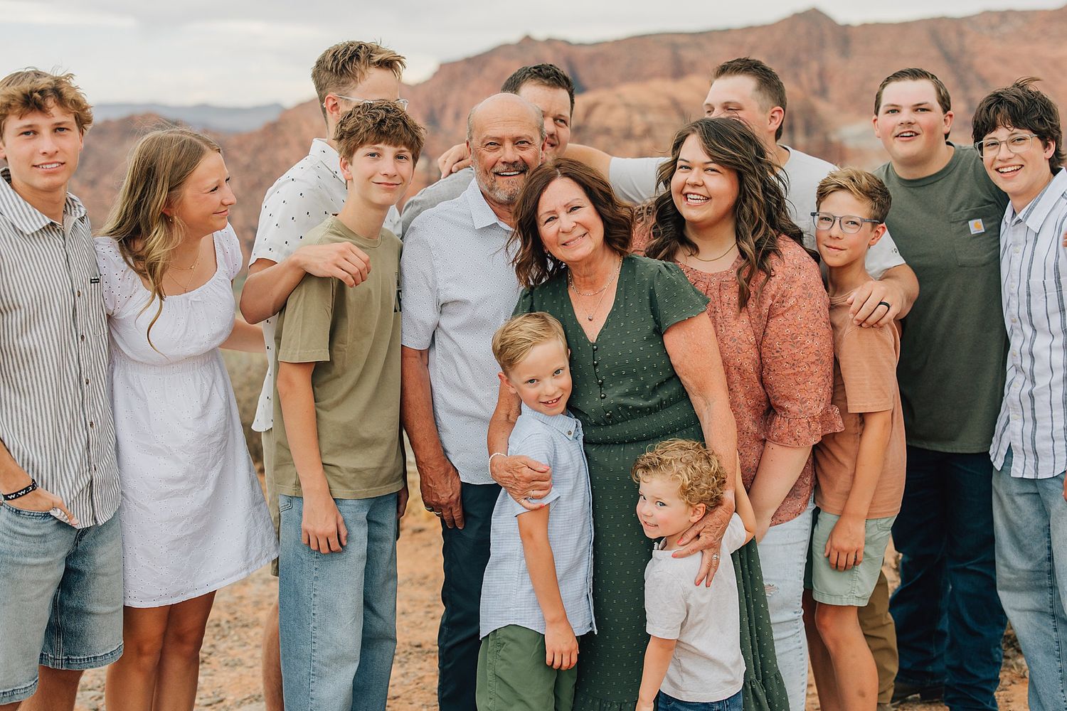 Large family group smiling together outdoors with red rock canyon landscape in background on sunny day.