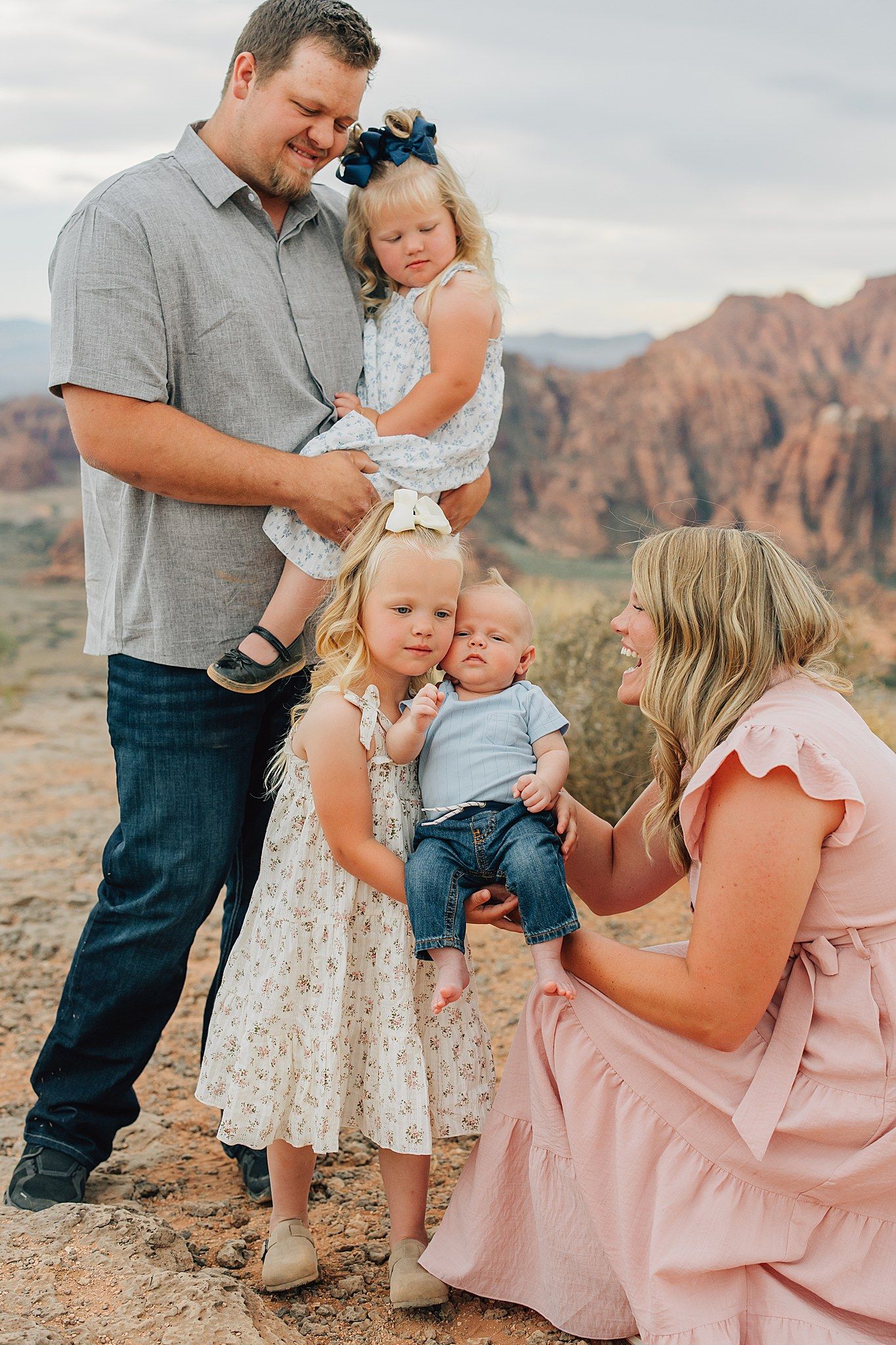 Family of five posing outdoors in red rock desert landscape, parents with three young children in coordinated outfits.