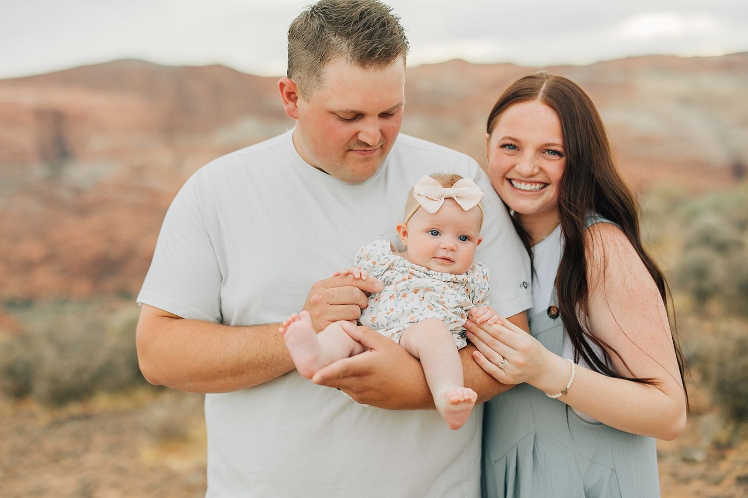 A happy young family with a mother, father, and baby girl posing outdoors in a scenic desert landscape.