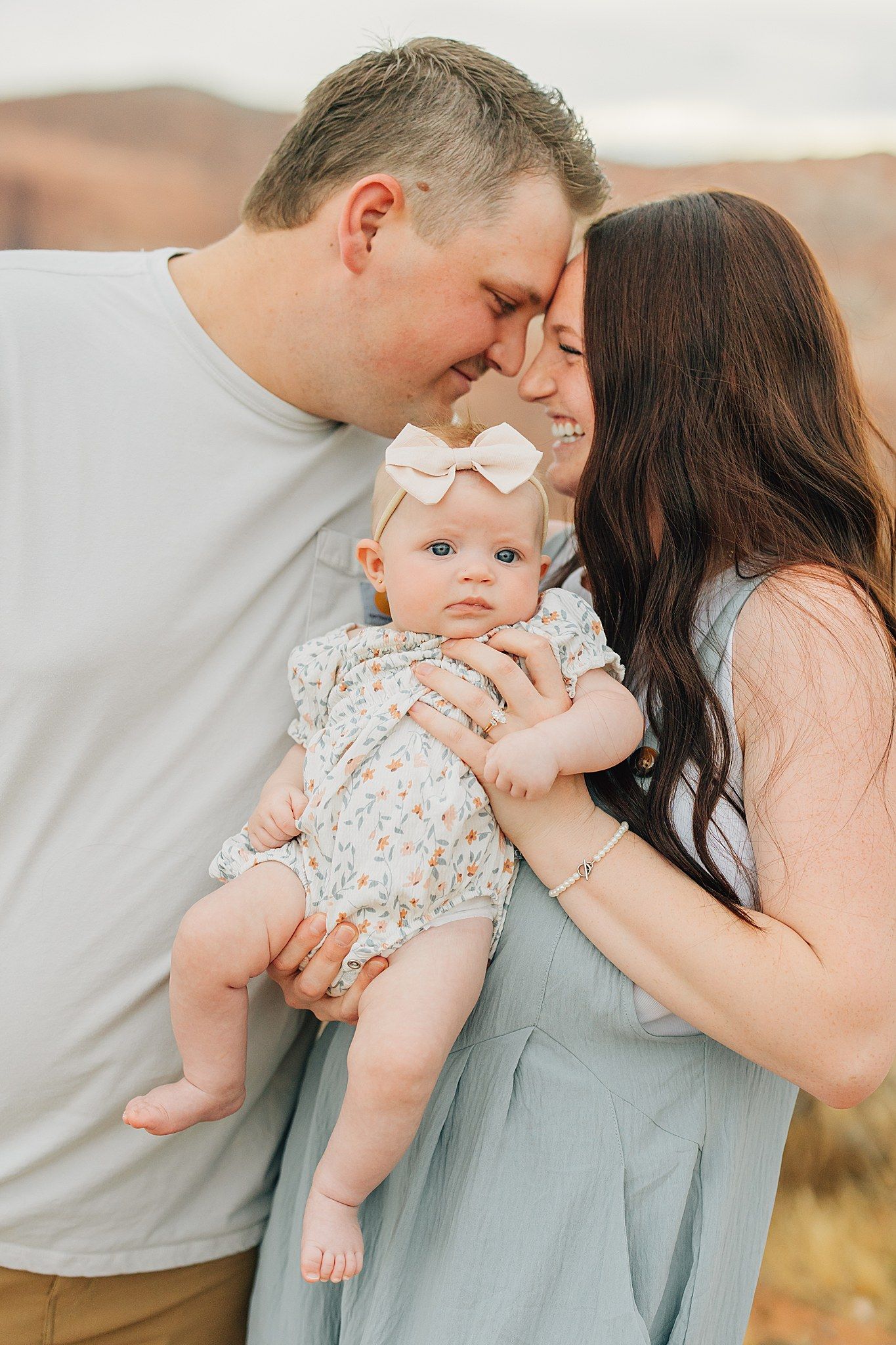 A couple tenderly touch foreheads while holding their baby girl in a floral romper and bow outdoors.