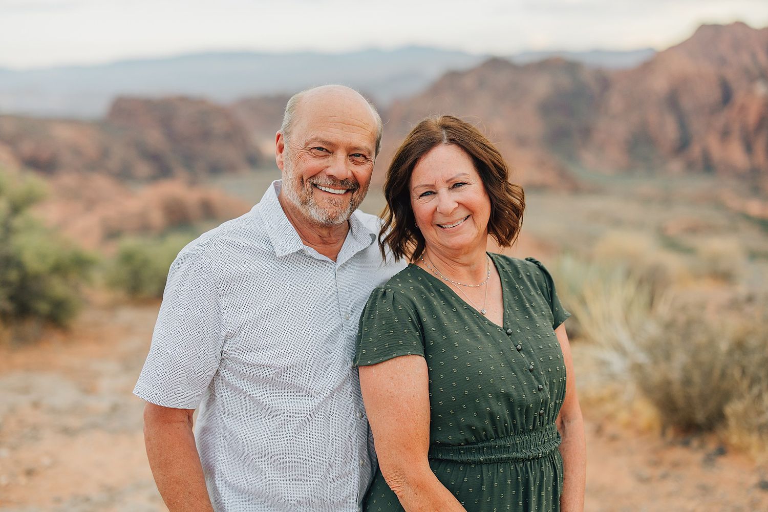 A smiling couple poses together outdoors with red rock canyon formations and desert landscape in the background.