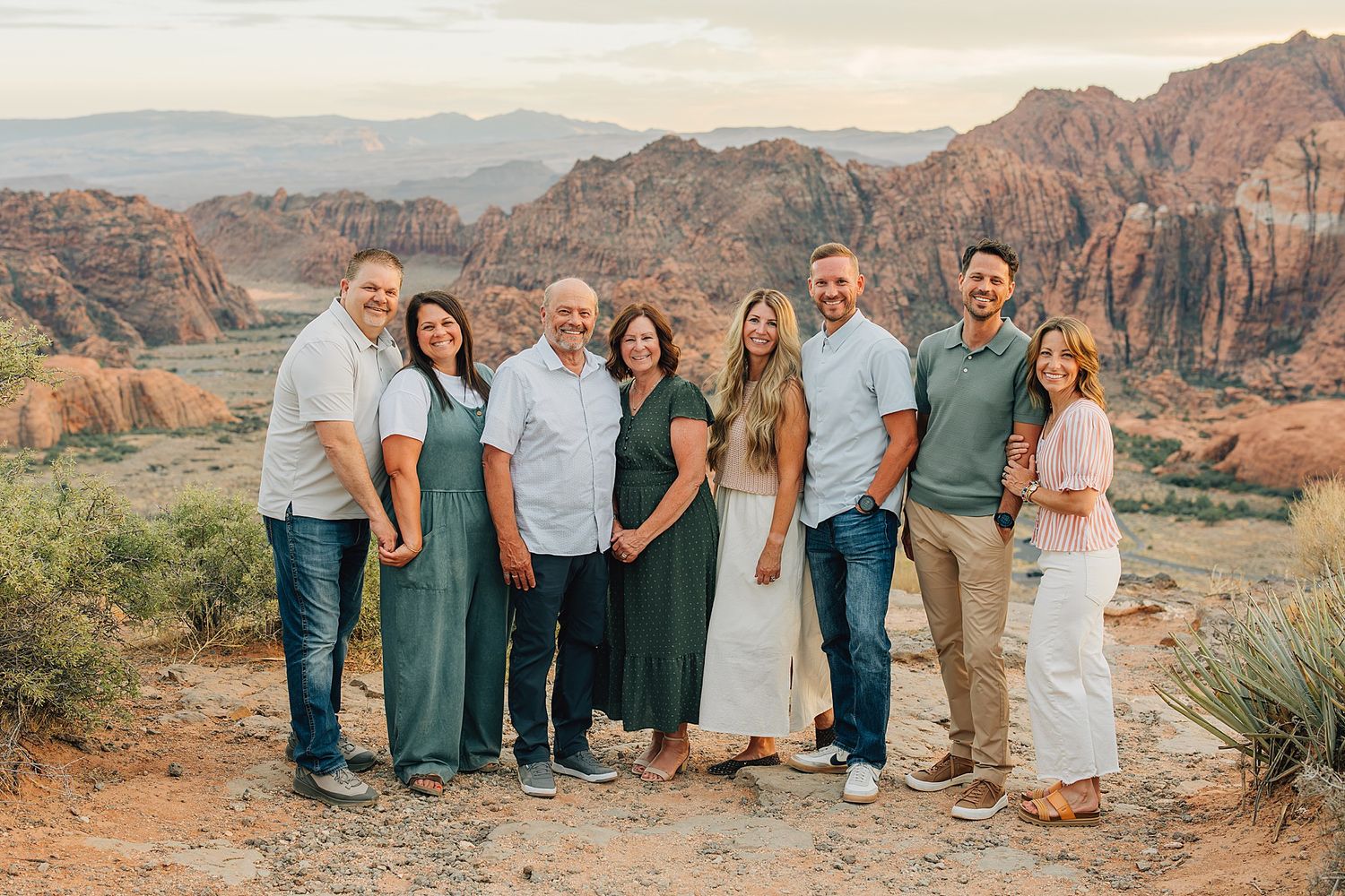 A joyful family group of eight poses together against a stunning red rock canyon landscape at sunset.