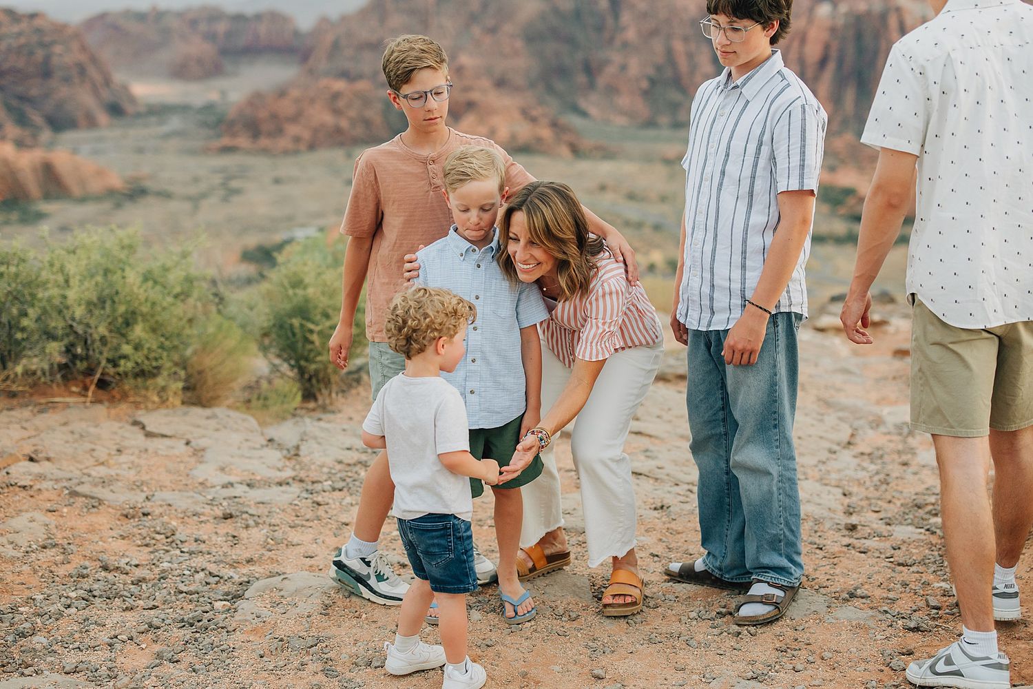 Family gathering outdoors in a desert landscape, mother bending down to young child surrounded by older boys.