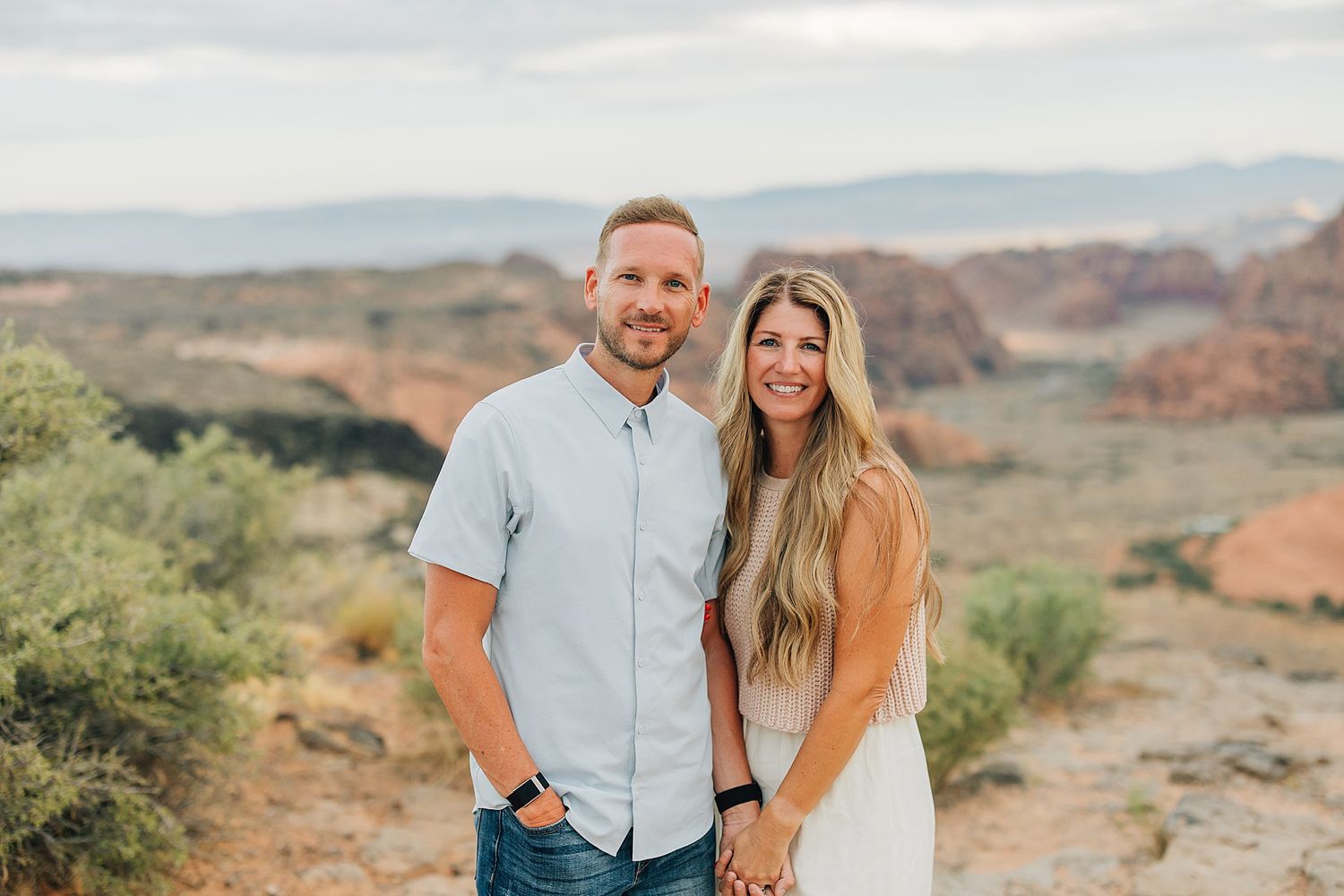 A smiling couple holds hands in a desert landscape with red rock formations and mountains in the background.