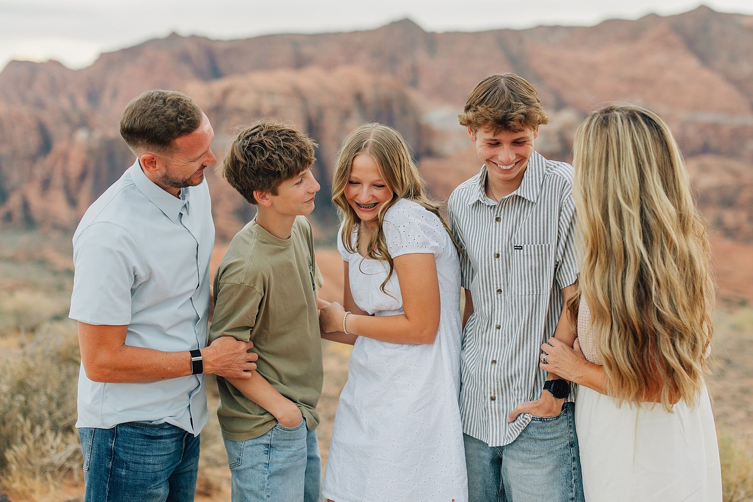 Family of five laughing together outdoors with red rock canyon landscape in background during golden hour.