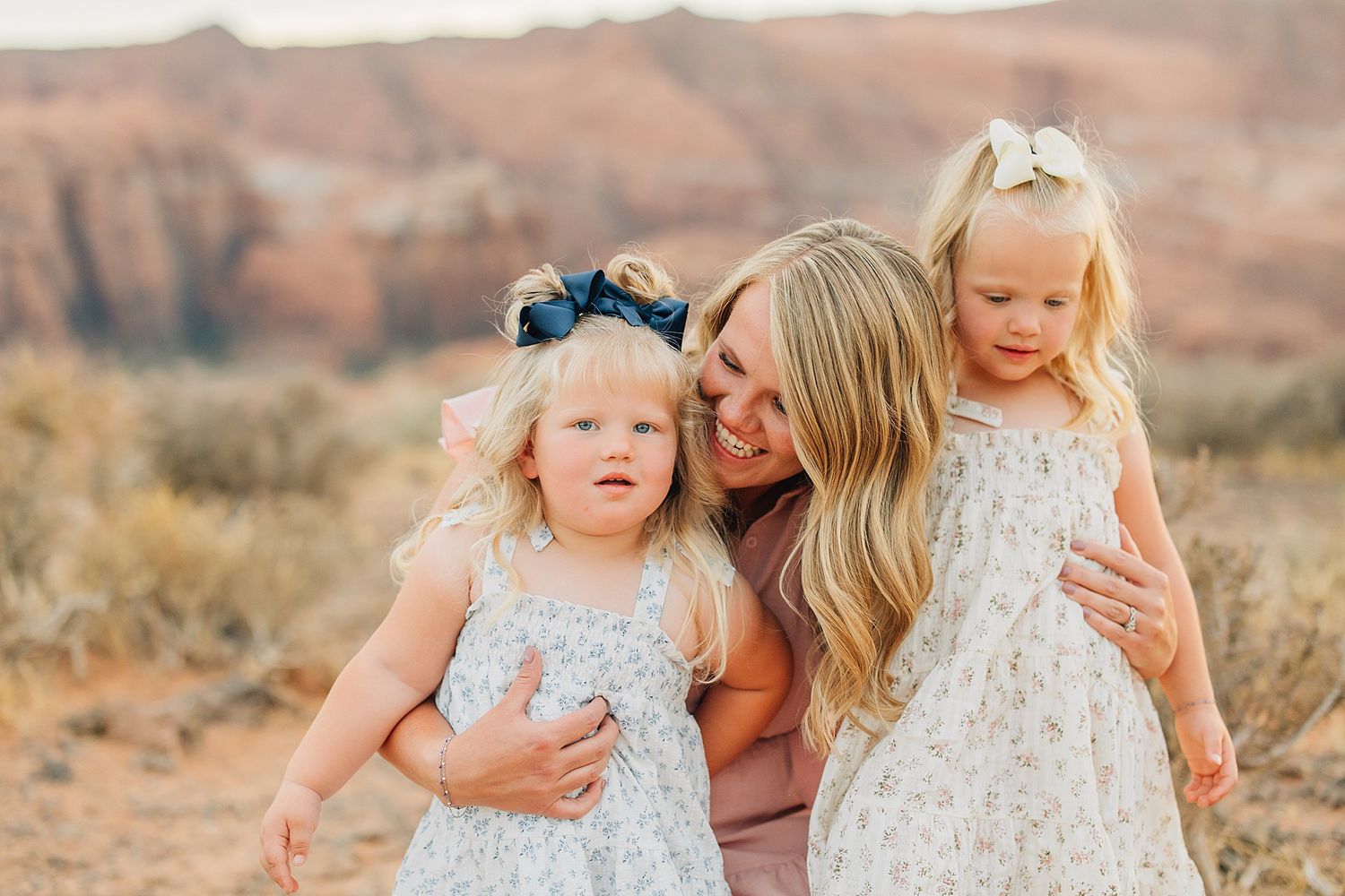 A smiling mother hugs her two young daughters in floral dresses with bows outdoors in a desert landscape.