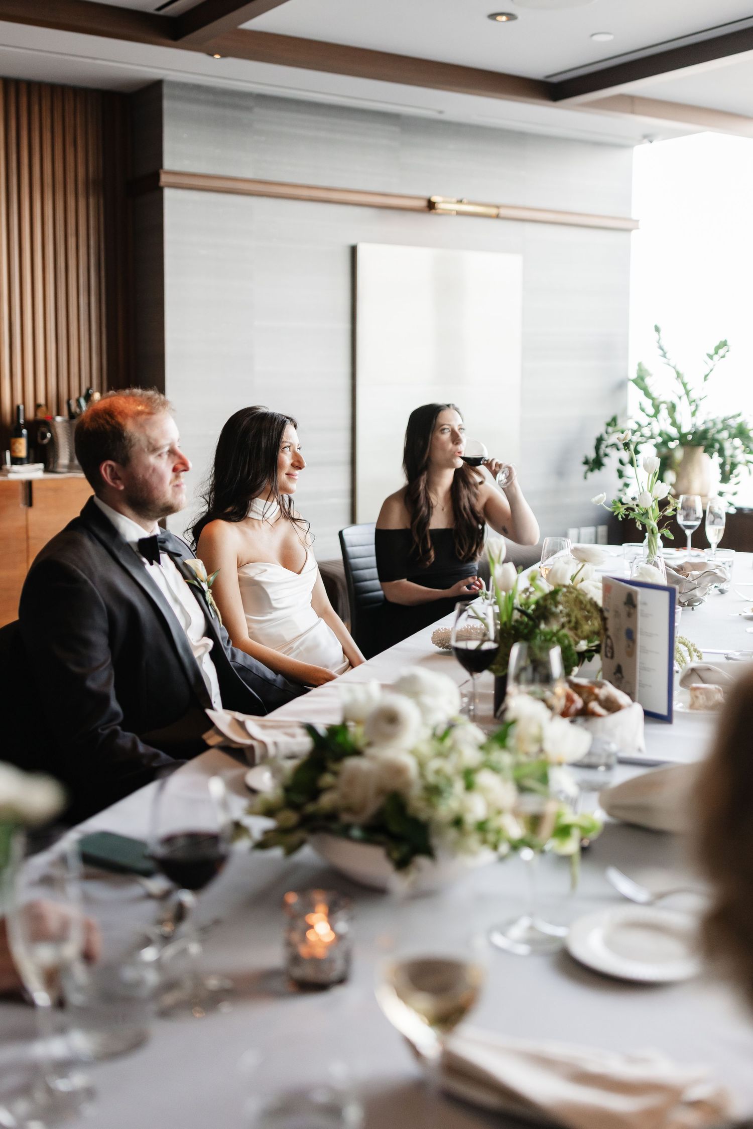 Bride and groom seated at elegant wedding reception table with floral centerpieces while bridesmaid gives a toast.
