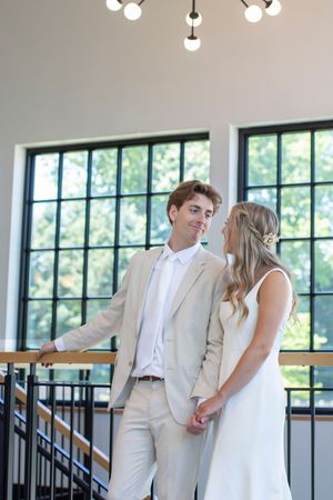 Bride and groom hold hands and share a smile near a staircase railing in a bright modern venue.