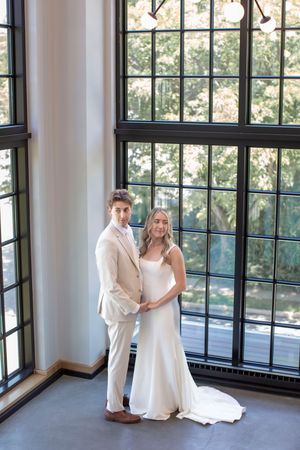 Bride in sleek white gown and groom in beige suit pose together before large industrial windows.