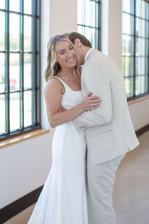 Smiling bride embraces groom as he kisses her cheek in a bright windowed wedding venue.