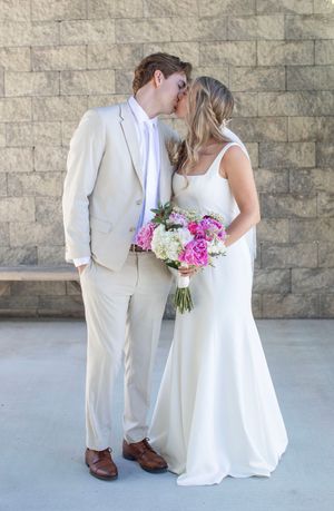 Bride and groom share a kiss while bride holds a vibrant pink and white floral bouquet outdoors.
