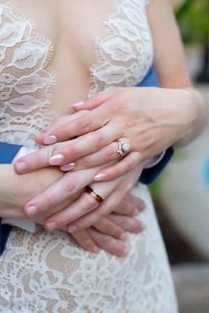 Close-up of newlyweds hands showing diamond engagement ring and red wedding band against lace gown.