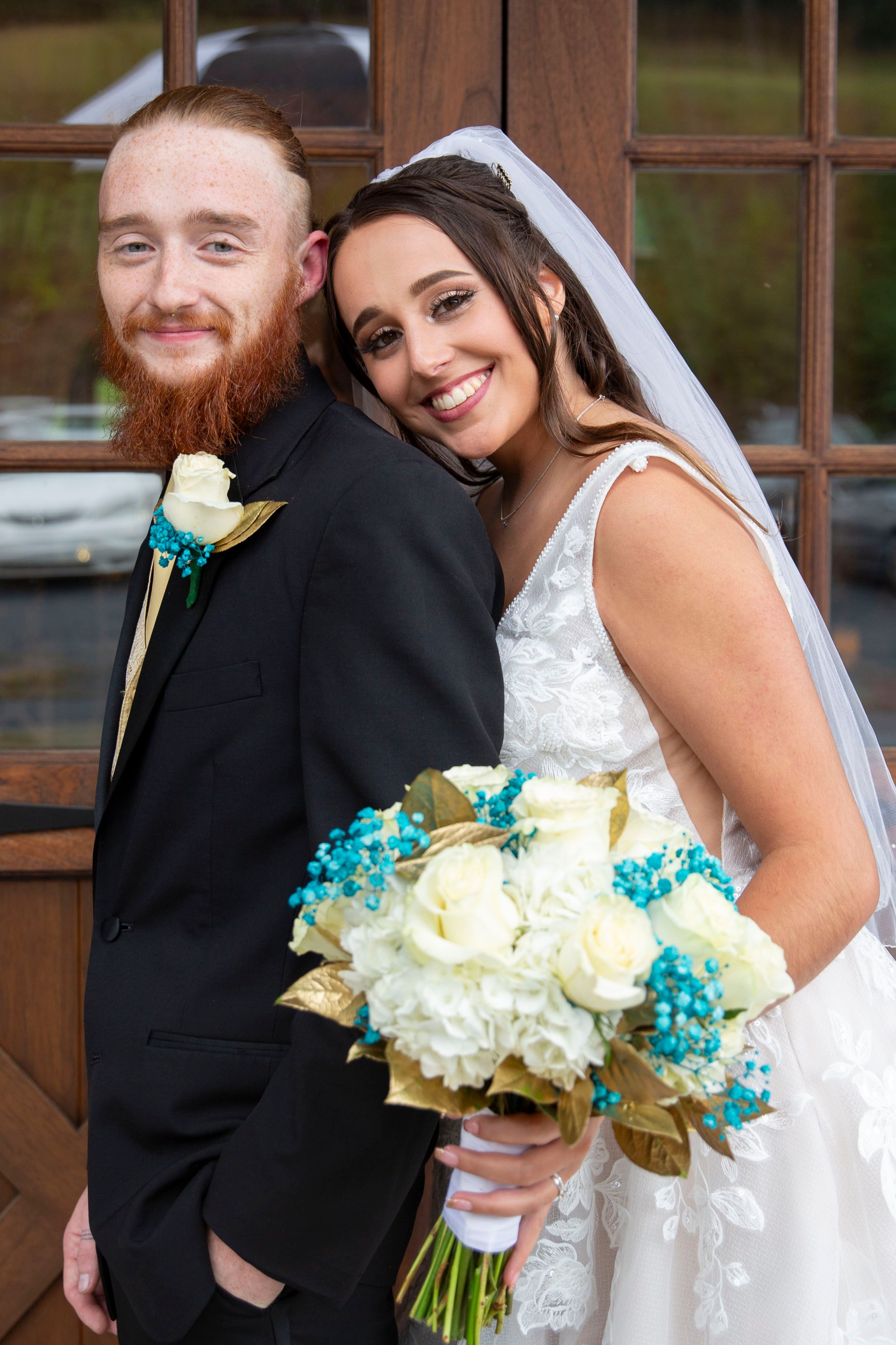 Smiling bride in lace gown holds white and teal bouquet beside red-bearded groom in black suit.