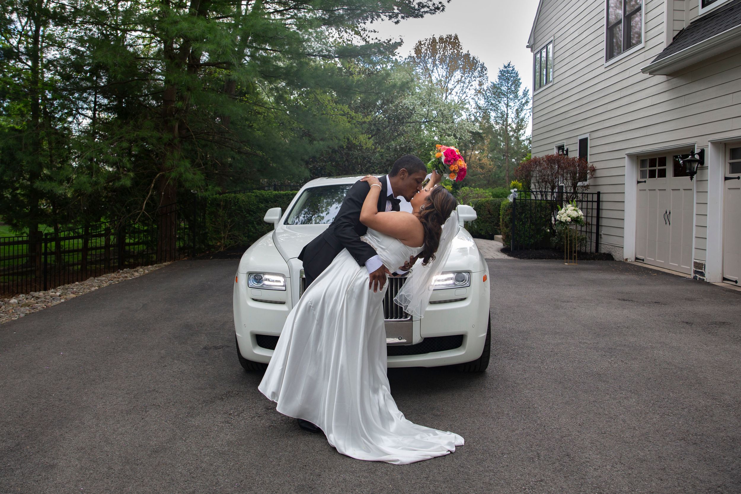 Bride and groom share a romantic dip kiss in front of a white Rolls-Royce luxury wedding car.