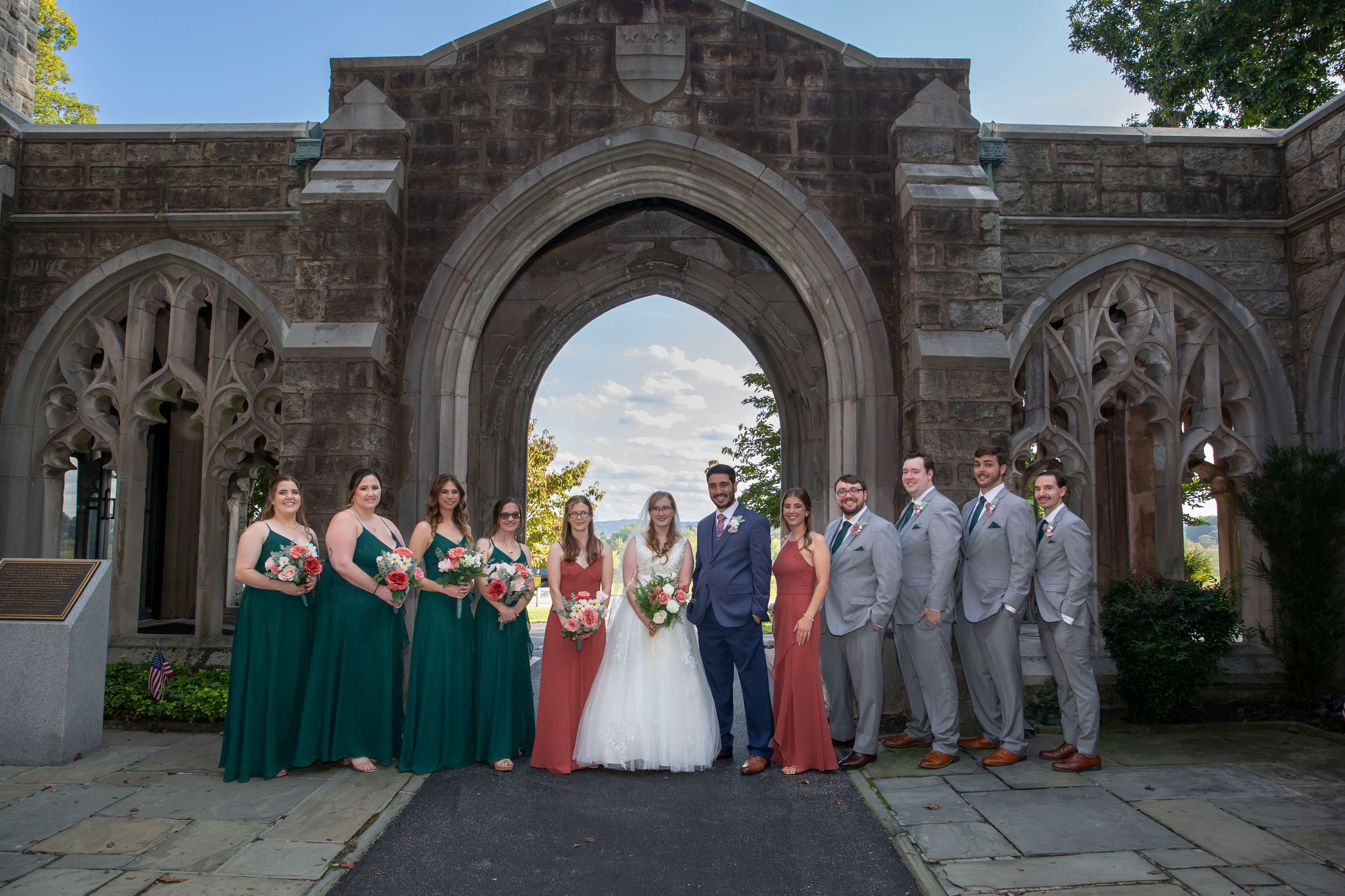 Wedding party poses in emerald green and gray attire beneath a stunning Gothic stone archway.