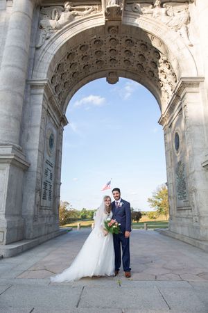 Bride and groom pose under a grand stone memorial arch on their wedding day.