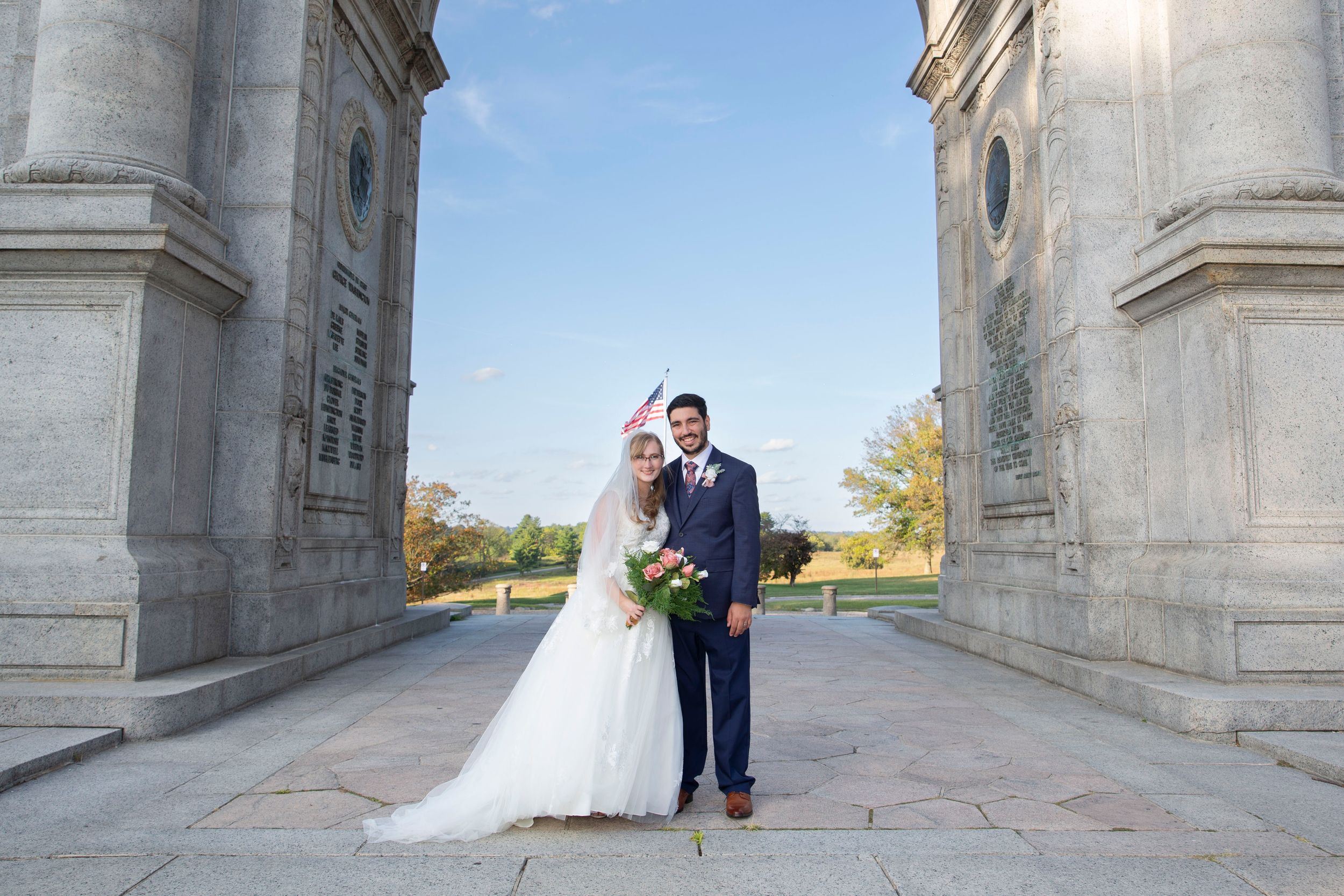 Bride and groom pose together between grand stone columns at an outdoor wedding venue on a sunny day.