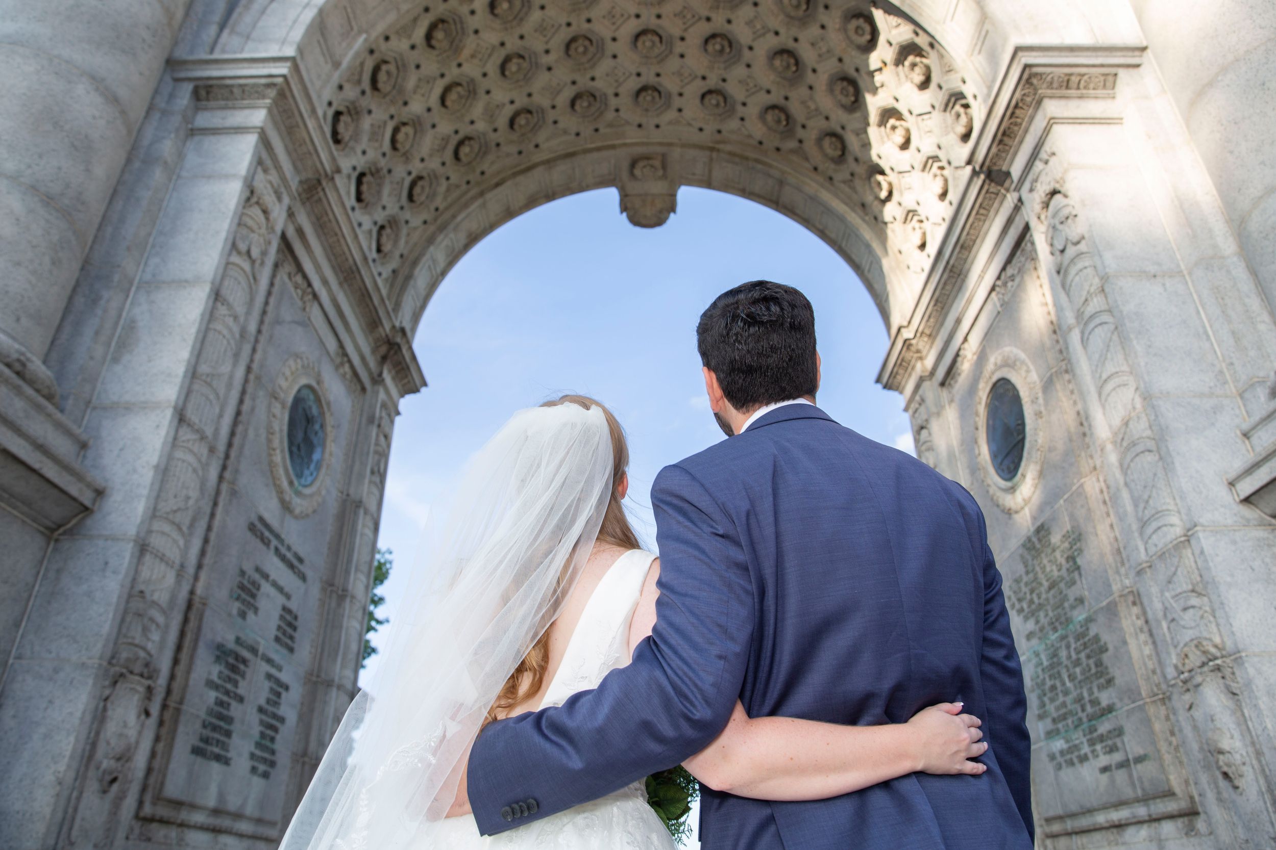 Bride and groom stand arm in arm facing a grand stone archway at their outdoor wedding venue.