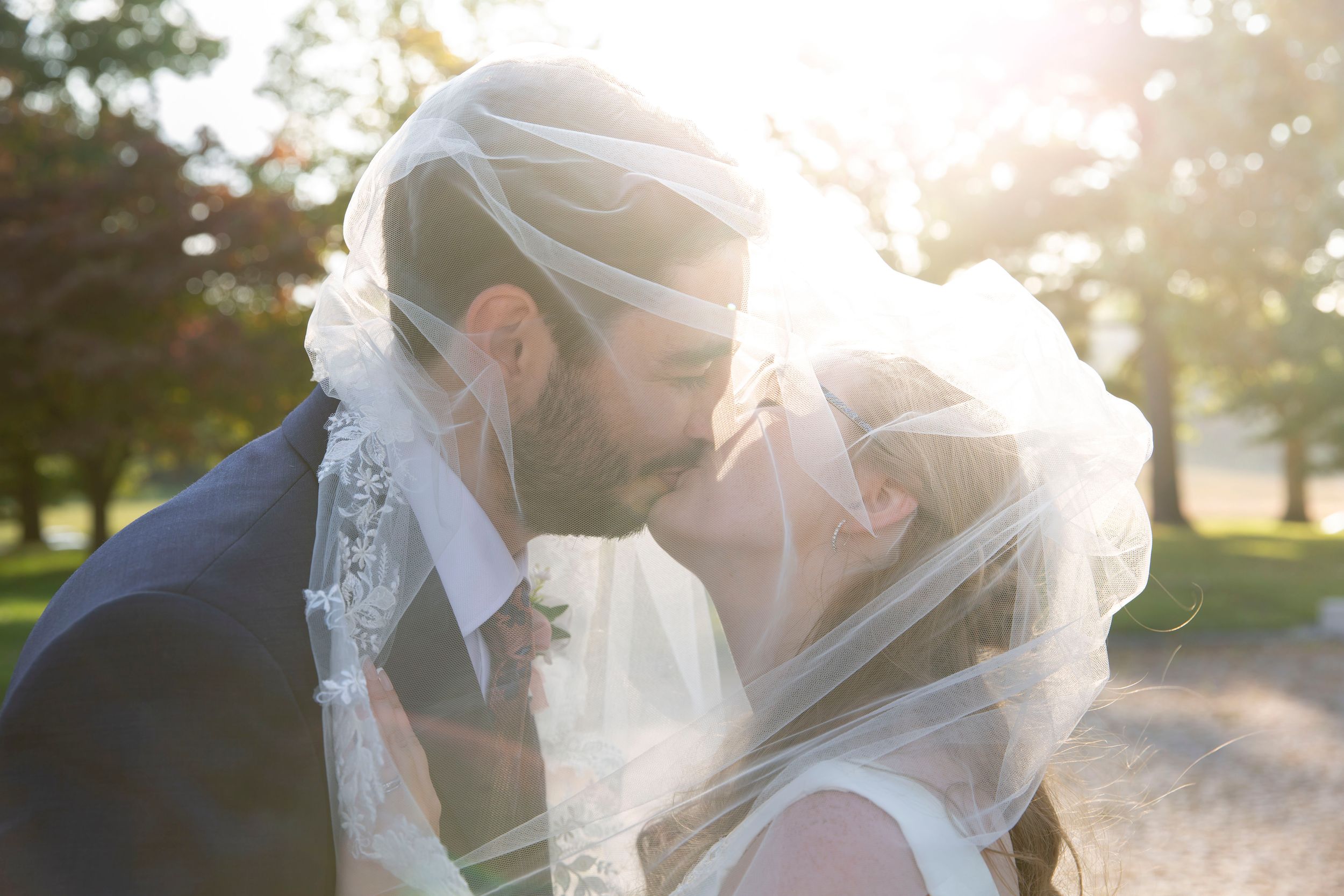 Bride and groom share a romantic kiss under a flowing veil with golden sunlight glowing behind them.
