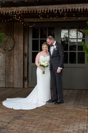 Elegant bride and groom pose together outside a rustic barn venue adorned with string lights.