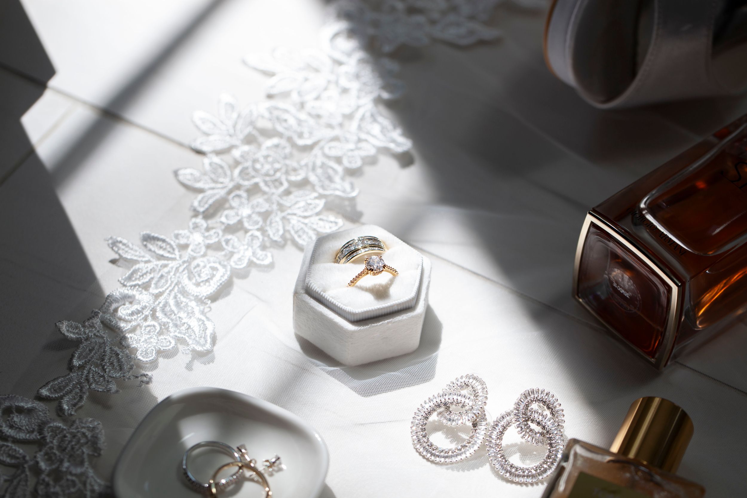 Elegant wedding flat lay featuring diamond earrings, gold rings, lace details, and perfume bottles.