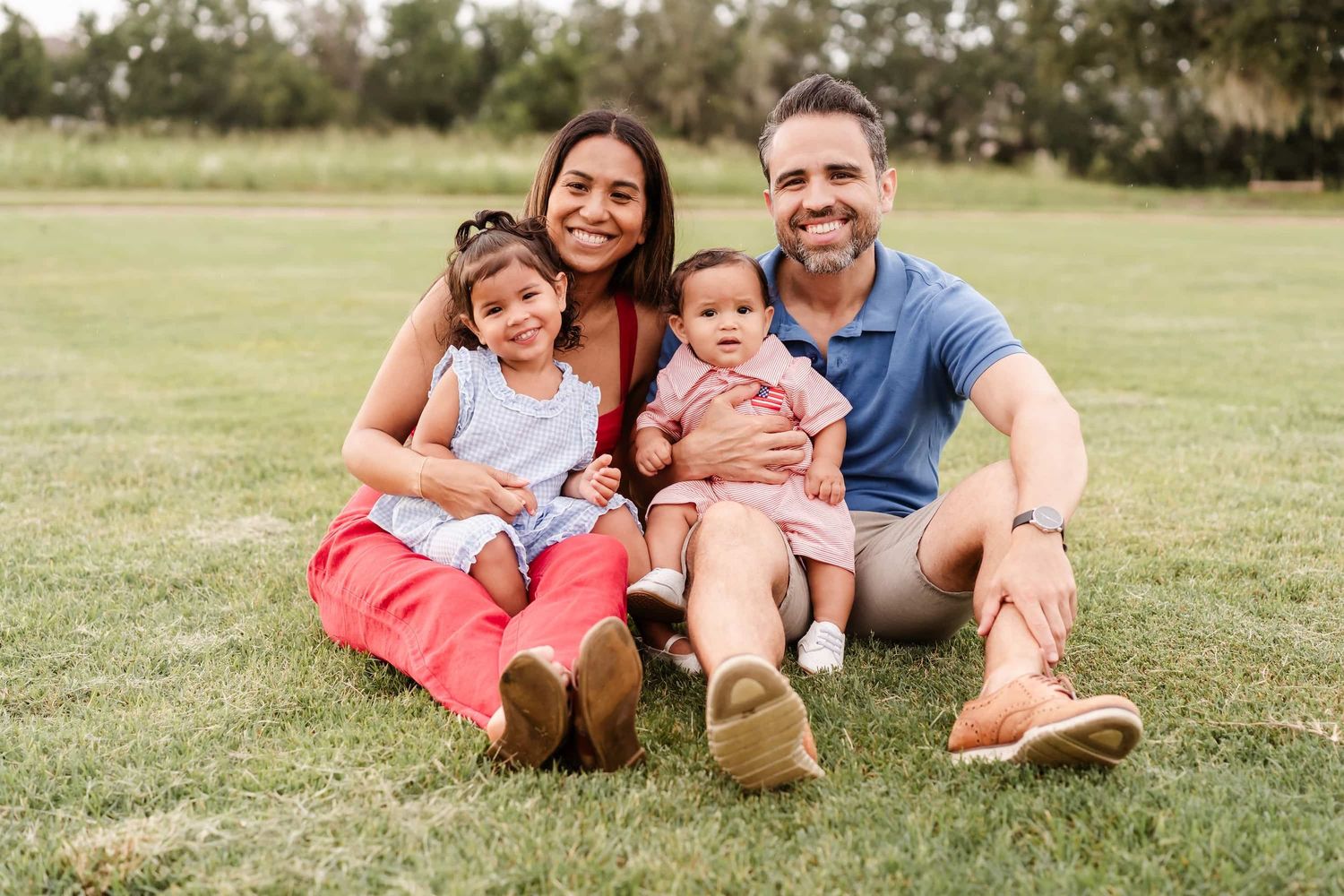 Parent in a red shirt lifts a giggling child wearing a red outfit up in the air at sunset in a grassy field.