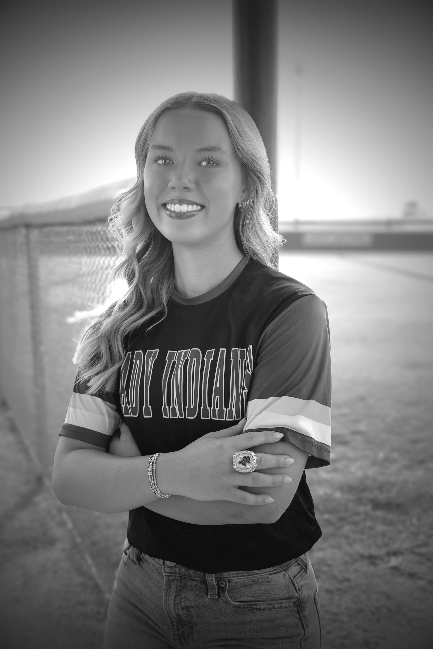 Black and white senior portrait of smiling blonde girl in Lady Indians jersey at baseball field.