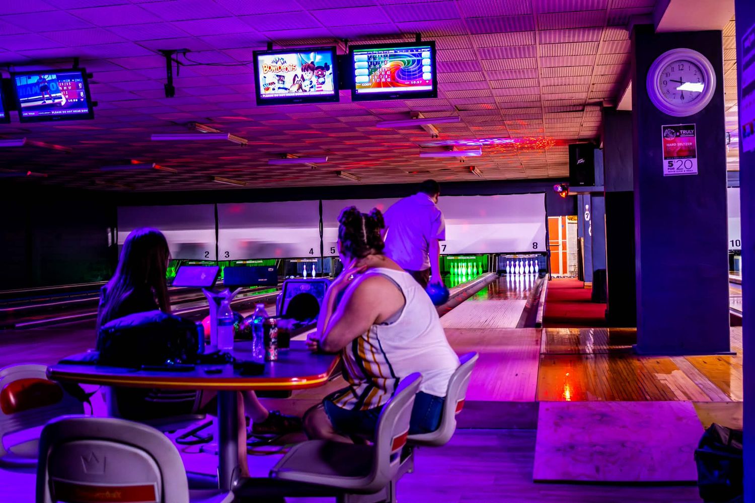People bowling at a vibrant neon-lit bowling alley with purple lights, TV screens, and colorful lanes in the background.