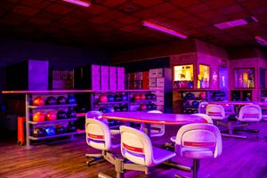 Bowling alley seating area with colorful balls on racks, white chairs, and vibrant purple neon lighting.