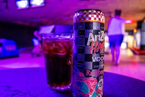 AriZona Hard Raspberry Vodka cocktail can and glass at a bowling alley with purple neon lighting.