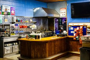 Interior of a casual fast food restaurant with wooden counter, commercial kitchen, stainless steel hood, and TV mounted on blue wall.