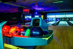 Colorful bowling balls on a ball return rack inside a neon-lit bowling alley with lanes in the background.
