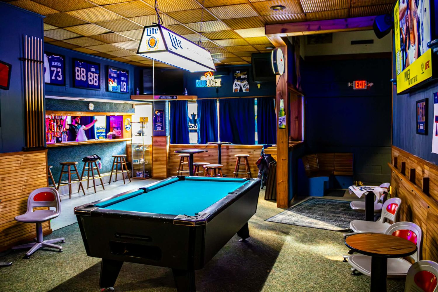 Sports bar interior with blue felt pool table, high-top seating, jerseys on walls, and a bar area in the background.