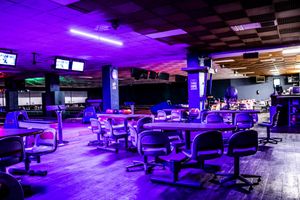 Empty bowling alley with purple neon lighting, round tables, and black chairs in a modern entertainment venue.