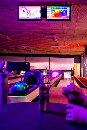 Colorful bowling alley with neon purple lights, bowling balls on return rack, pins at end of lanes, and overhead TV screens.