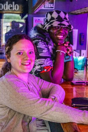 Two friends smile at a bar with purple lighting, Modelo sign visible in background.