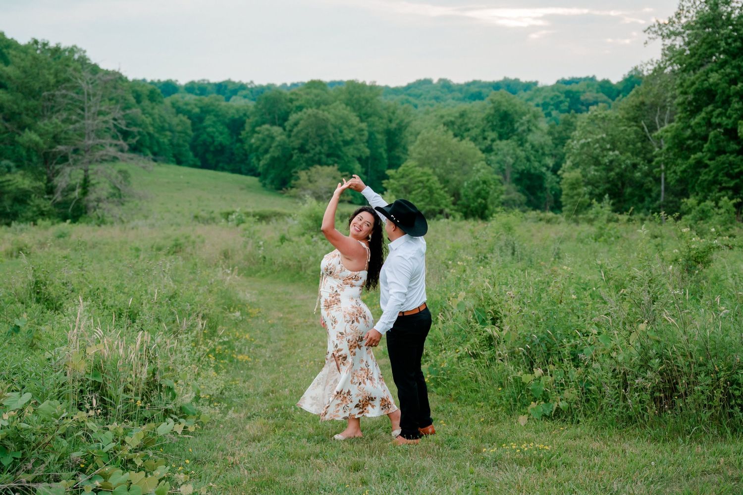 Couple dancing together in a lush green meadow, woman in floral dress being twirled by partner at golden hour.