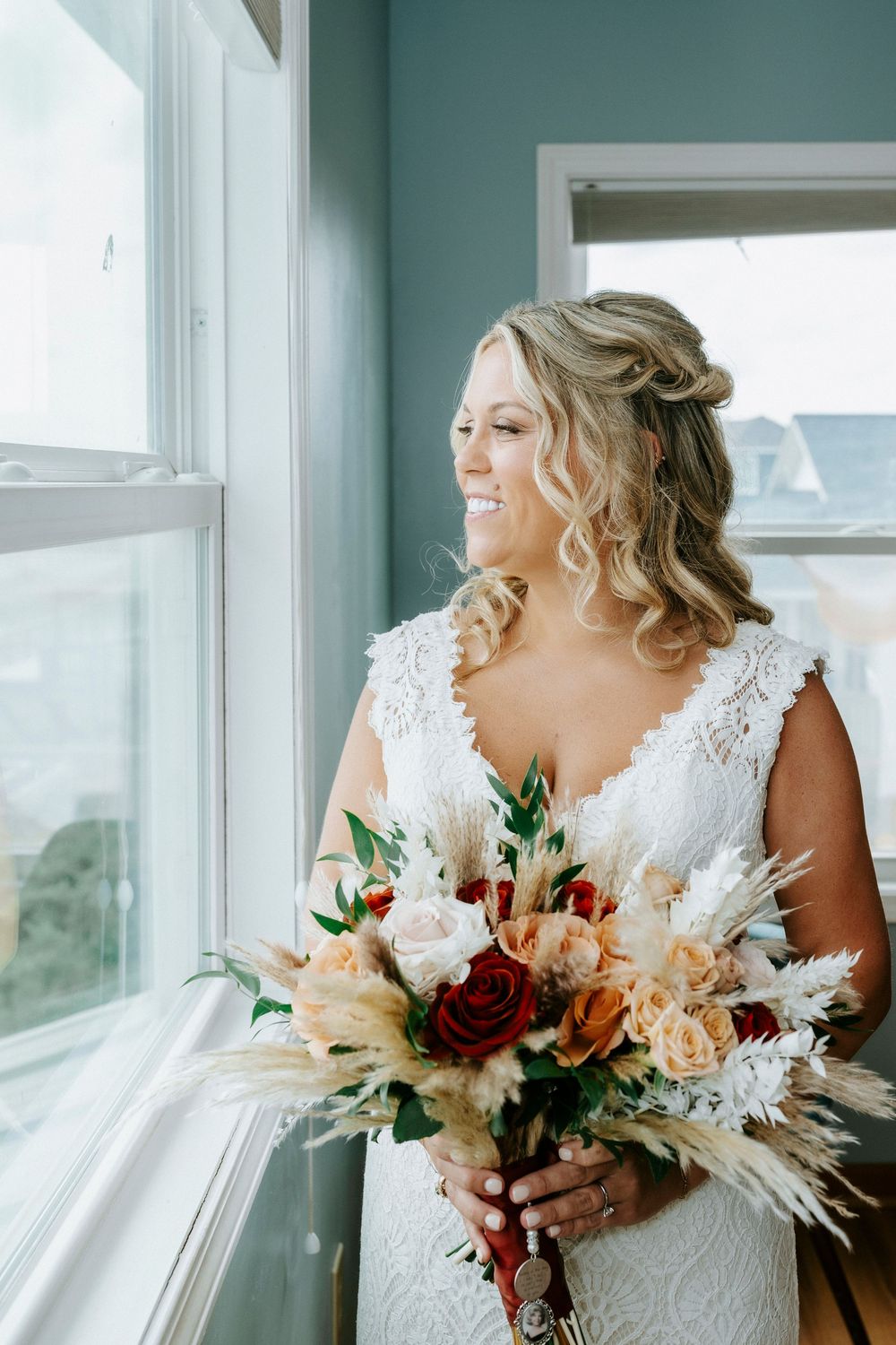 Smiling bride in lace gown holds boho bouquet of roses and pampas grass by a sunlit window.