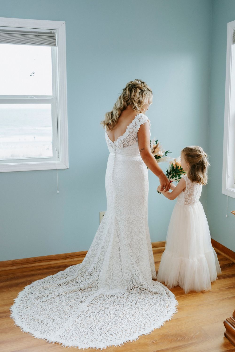 Bride in lace wedding gown holds hands with flower girl in white dress, standing by ocean-view window.