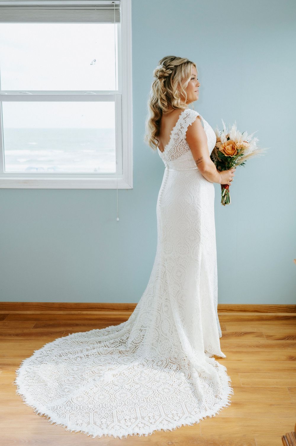 Bride in white lace wedding gown with train holds bouquet, standing by ocean-view window.